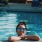 woman with sunglasses, in a pool, rests her arms on the ledge and smiles